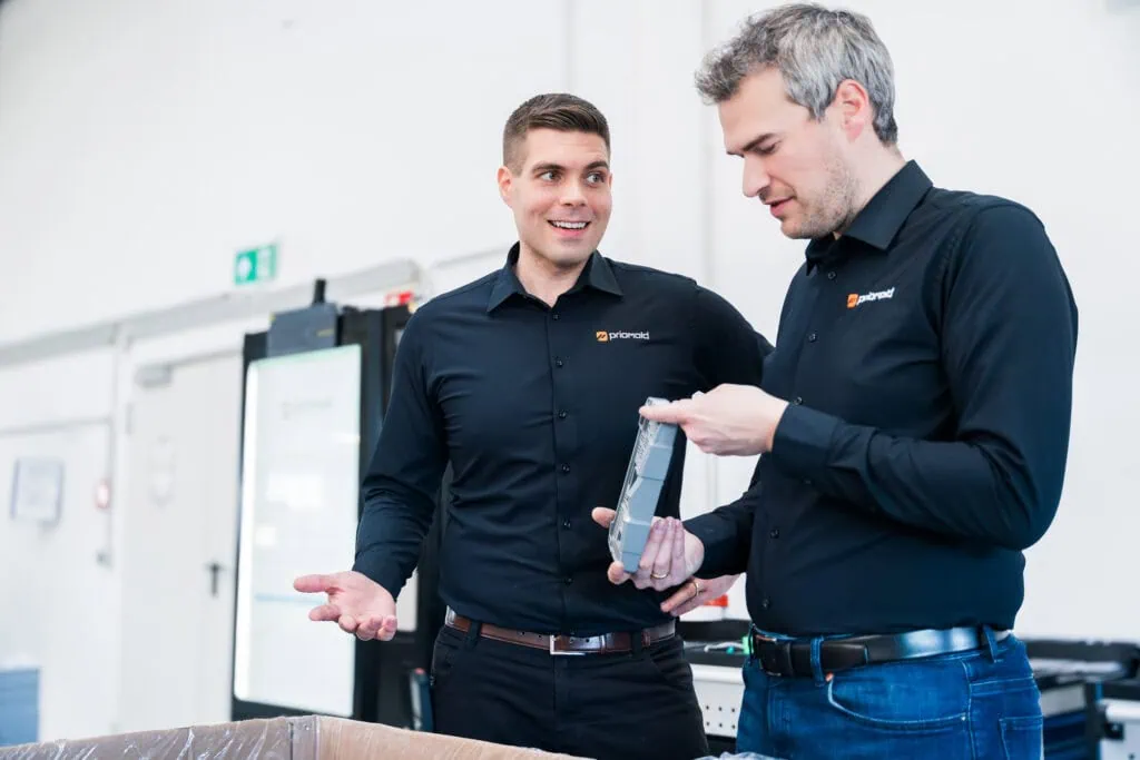 Die priomold-Geschäftsführer Thomas Schönbucher und Moritz Zumdick in der Produktion bei priomold mit einem Kunststoffbauteil als Symbol für Spritzguss-Kompetenz und kurze Wege.